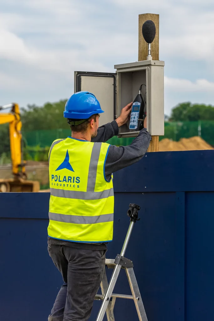 polaris acoustics acoustic consultants for planning applications setting up a construction noise monitoring sound level meter at site hoarding on a step ladder and PPE. Noise assessments.