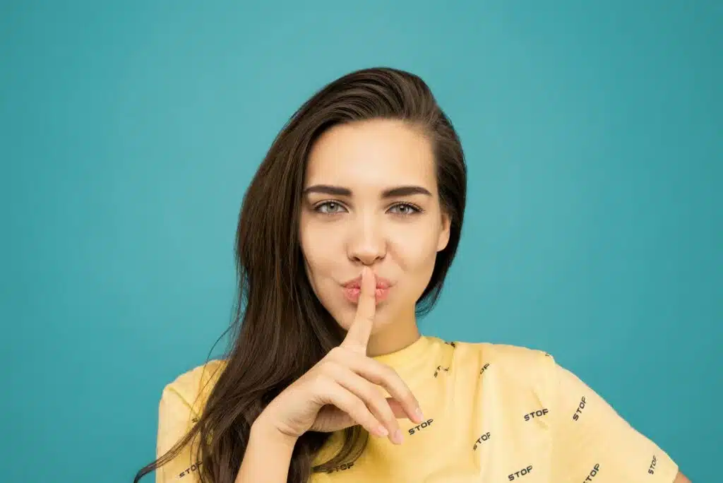 Portrait of a woman with brown hair posing with a hush gesture against a blue background. are air source heat pumps noisy? 7 proven facts.