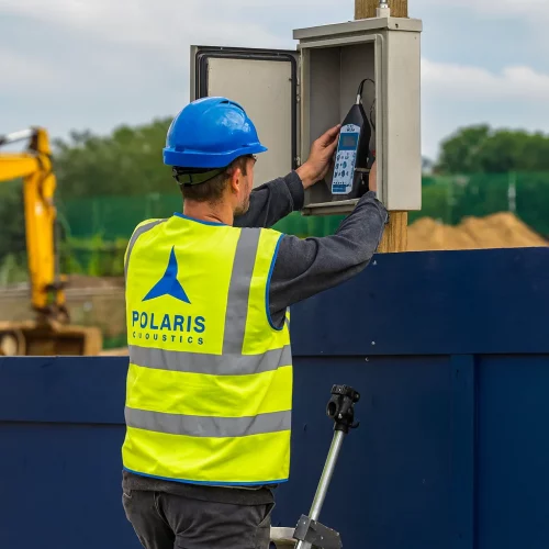 polaris acoustics acoustic consultants for planning applications setting up a construction noise monitoring sound level meter at site hoarding on a step ladder and PPE. Noise assessments.
